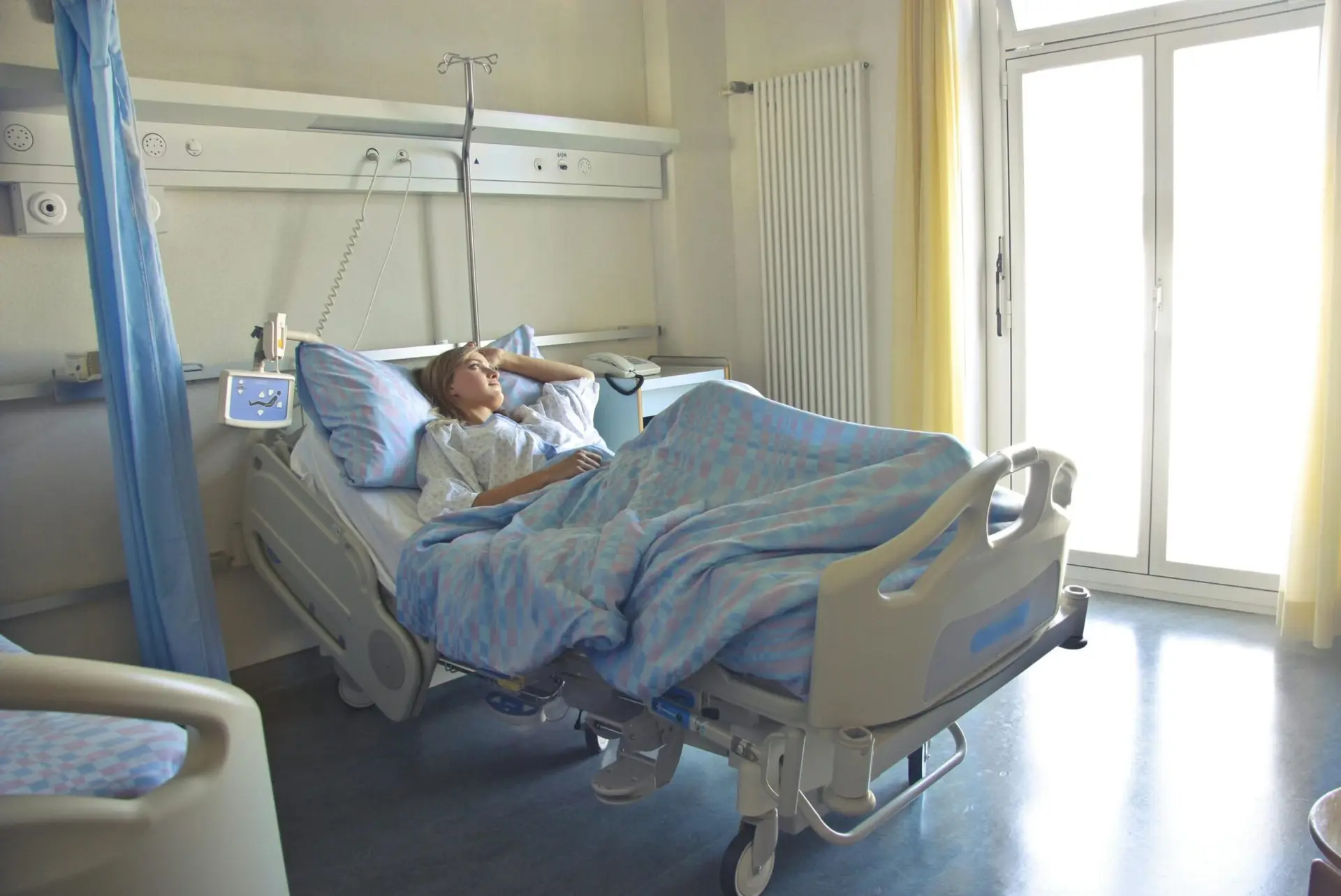 A woman lying in a hospital bed in a bright, softly lit room, appearing to rest or recover.